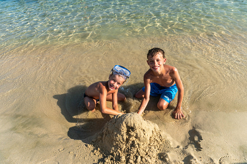 portrait of two happy boys, on the beach, on the sand, playing with the sand, making something out of the sand, on the seashore, in swimwear