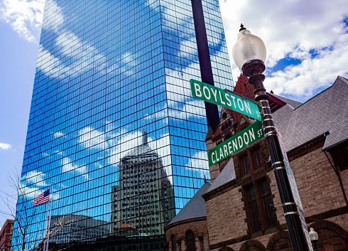 Boston, Massachusetts, USA - April 16, 2024: Close-up of Boylston and Clarendon street signs on a street light pole in Copley Square, a public square in Boston's Back Bay neighborhood. In the background is the 200 Clarendon Street skyscraper, formerly named the John Hancock Tower, and a small portion of Trinity Church. The reflection of clouds and the old John Hancock building is in the blue glass of the Hancock Tower. The Hancock Tower is highest building in Boston and all of New England.