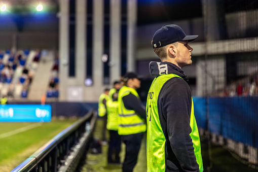 Male security professional in a high-visibility vest standing guard at an outdoor football stadium during an evening event, maintaining crowd safety and venue security.