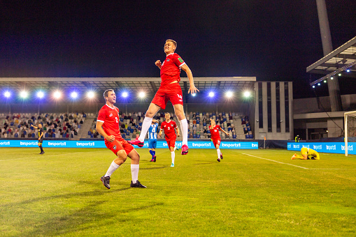 An energetic male soccer player jumps to celebrate a goal during a night football match. The professional athlete is wearing a red uniform, and the floodlit stadium creates a dynamic backdrop. Soccer game action captured outdoors.