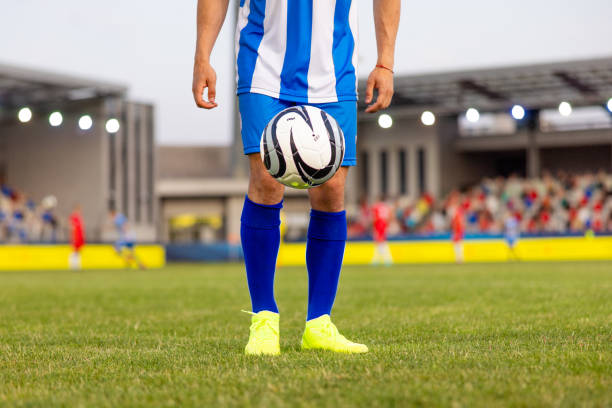 Unrecognizable male soccer player in striped blue and white uniform standing with a black and white ball on the grassy field, neon cleats visible, stadium audience in the background, outdoors.