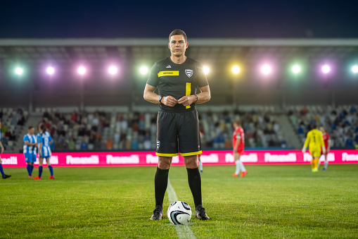 A confident male Caucasian referee stands on the pitch with a ball at his feet. He is dressed in a professional sports uniform, positioned under stadium lights with teammates in the background.