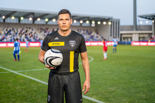 A male referee in professional black and yellow attire holds a soccer ball on the football pitch, with players and stadium in the background, conveying authority and focus during a match.