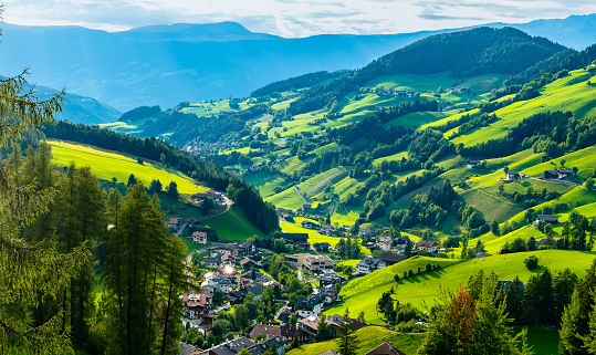 A small town nestled in the centre of a lush green valley, surrounded by rolling hills and mountains. The landscape is marked by vibrant green fields, patches of forests, and winding roads connecting homes and farms. In the distance, layers of mountains fade into the horizon under a clear blue sky. Italian Alps.