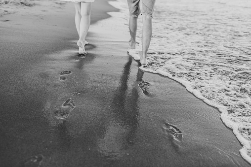 Down view of bare feet on sand beach. Couple in love walking barefoot on water with big waves on seashore. Man and woman walk on sea. Time together. Bottom view on legs. Back view. Black white photo