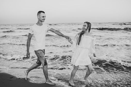 Black and white photo. Happy couple in love holding hands and looking at each other walking on water with big waves on seashore. Man and woman walk on beach sea. Spending time together
