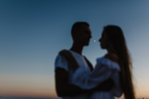 Blurred silhouette of a man hugging a woman at sunset against the background of moon in the sky. Female embrace male and enjoy evening on summer day. Couple in love on seashore in sand sea.