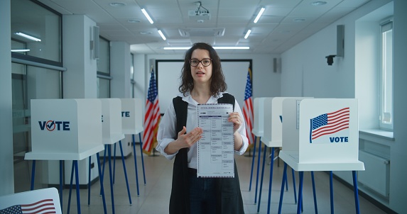 American female voter or polling officer speaks on camera, shows paper ballot, calls for voting. National Election Day in the United States. Voting booths at polling station. Civic duty and patriotism