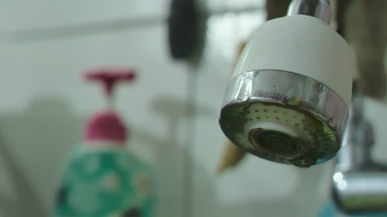 Close up of the sink faucet leaking and dripping water in the dirty and dark kitchen, depicting a messy and neglected home