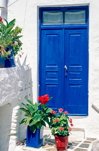 Blue-painted double door of a white-tiled house or guesthouse with flower pots as decoration on the Greek island of San Turin in the afternoon sunlight