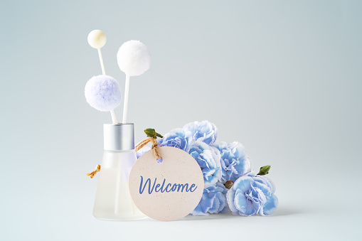 Welcome paper tag with fragrance diffuser and flowers on vintage blue background, copy space