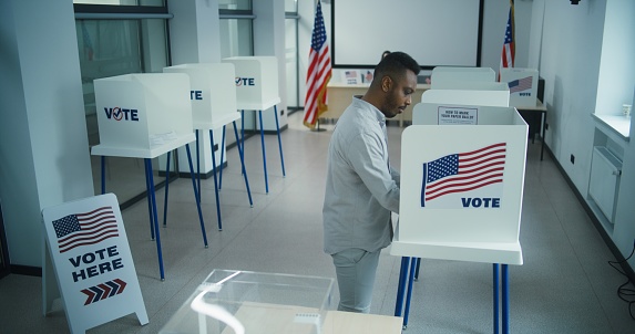 African American man votes for presidential candidate in voting booth at polling station, then puts paper ballot in box. Male US citizen during National Elections Day in the United States. Civic duty.