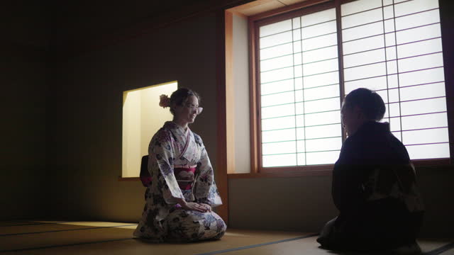 Female caucasian tourist in kimono learning manners of greetings from a senior instructor in dark Japanese tatami room
