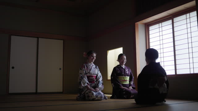 Female tourists in kimono learning manners of greetings from a senior instructor in dark Japanese tatami room - smiling
