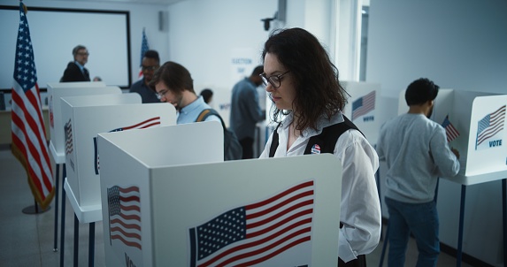 Woman in glasses votes in booth in polling station office. National Election Day in United States. Political races of US presidential candidates. Civic duty and patriotism. Slow motion. Dolly shot.