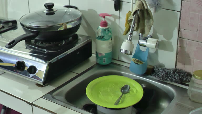 The sink faucet is leaking and dripping water in the dirty and dark kitchen, dirty dishes and pans have not been washed, depicting a messy and unkempt house