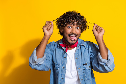 Photo portrait of nice young male hold curly strands stick tongue wear trendy denim outfit red scarf isolated on yellow color background.