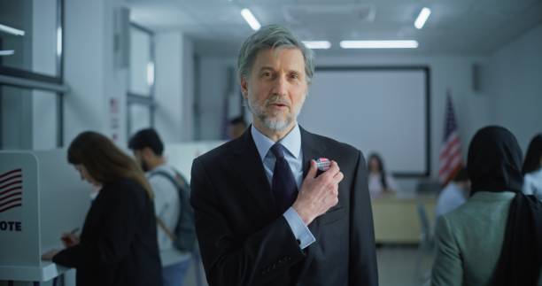 Portrait of man, United States of America elections voter. Businessman with badge speaks on camera and calls for voting in modern polling station. Background with voting booths. Concept of civic duty.