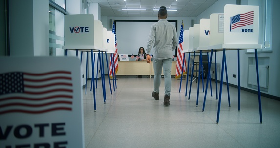 US citizen, African American male voter walks to female polling officer for registration at polling station, takes paper bulletin for voting. National Election Day in the United States of America.