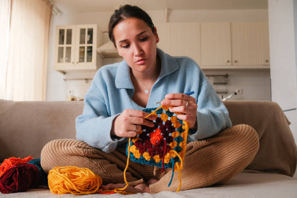 istock Young woman knitting crocheting with colored yarn granny square while watching online tutorial on phone at home. Woman watching needlework lessons, home hobbies 2149124818