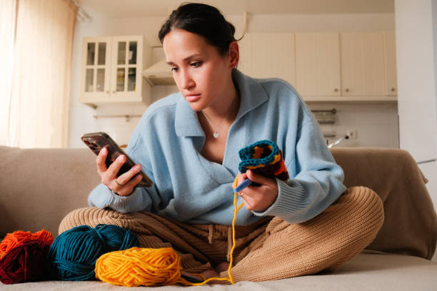 istock Young woman knitting crocheting with colored yarn granny square while watching online tutorial on phone at home. Woman watching needlework lessons, home hobbies 2149125204