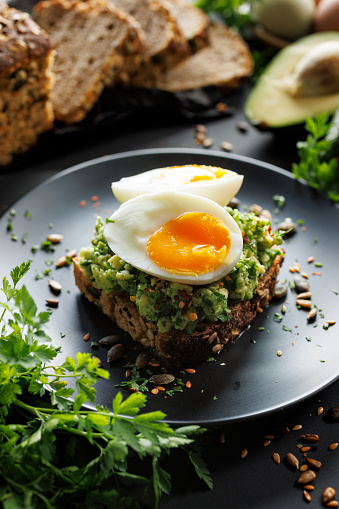Avocado sandwich made of sourdough rye bread with mashed avocado, soft-boiled egg and fresh herbs on a black plate, close up view