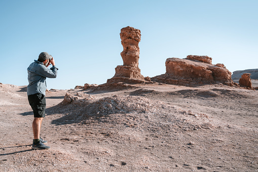 A male photographer taking pictures of a distinctive rock sculpture in the Atacama Desert, Chile, with clear blue skies and rugged terrain around.