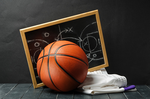 A basketball rests against a blackboard with tactical diagrams chalked on it, symbolizing a coachs strategic planning. The blackboard is in a wooden frame, and a white towel and colored chalks lie on the dark floor beside the ball.