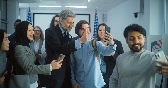 Happy multiethnic US citizens take selfies with mature presidential candidate after voting. Diverse people with politician at polling station. National Election Day in the United States. Slow motion.