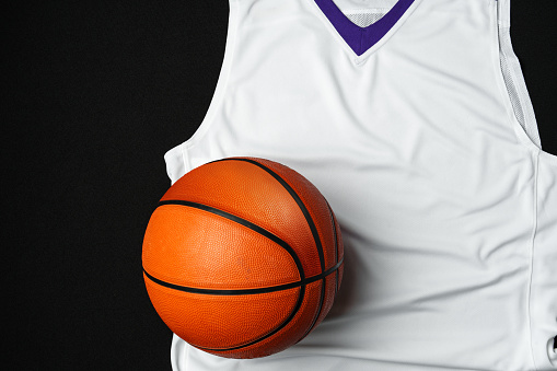 A basketball rests against a white and purple basketball jersey with a v-neck collar on a contrasting black backdrop, highlighting the textures and details of the sports equipment and apparel commonly used in basketball games.