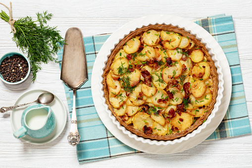 Irish potato pie of crispy crust layered with potatoes, bacon and onion in baking dish on white wooden table with ingredients and cake shovel, horizontal view from above, flat lay
