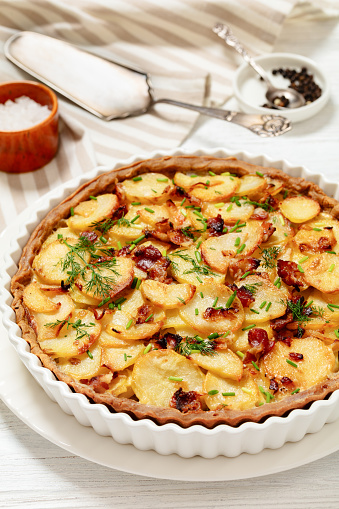 Irish potato pie of crispy crust layered with potatoes, bacon and onion in baking dish on white wooden table, vertical view from above, close-up
