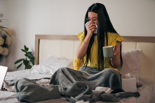 A young sick woman spends the weekend at home, lying in bed covered with a blanket and drinking a healthy warm drink.