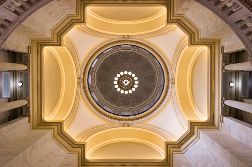 A large round chandelier with two tiers, segmented into six sections, the bottom tier holds 12 bulbs and the upper has 6. It hangs from an an ornately decorated ceiling with intricate coving and design.