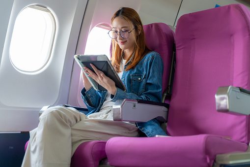 Happy young woman using her tablet while sitting alone on the plane by the window