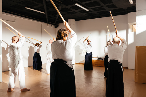 Students clad in white uniforms stand in rows inside a dojo, diligently practicing wooden sword techniques under the guidance of their instructor.