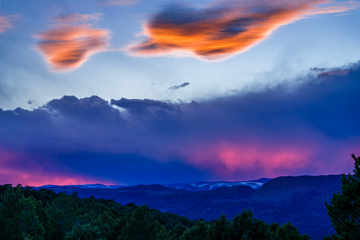 Dramatic Colorful Mountain View at Sunset - Rugged peaks, valleys and forests in scenic mountainous landscape. Colorful pink light filtering through clouds while upper clouds being lit with warm rays of sunlight.