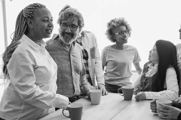 Multiracial friends with different ages drinking coffee together indoor - Multi generational people during tea break inside coworking office kitchen - Black and white editing