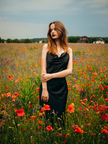 Beautiful young girl in a black evening dress posing against a poppy field on a cloudy summer day. Portrait of a female model outdoors. Rainy weather. Gray clouds.