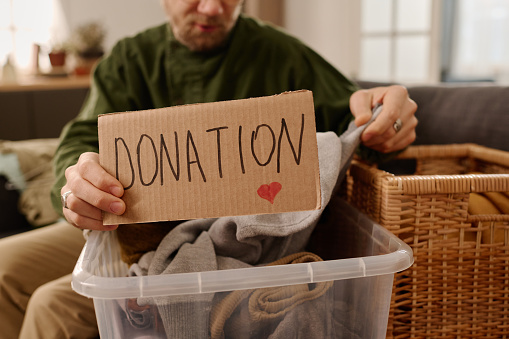 Young man or volunteer holding piece of cardboard with word donation over plastic box with heap of clothes while choosing attire for charity