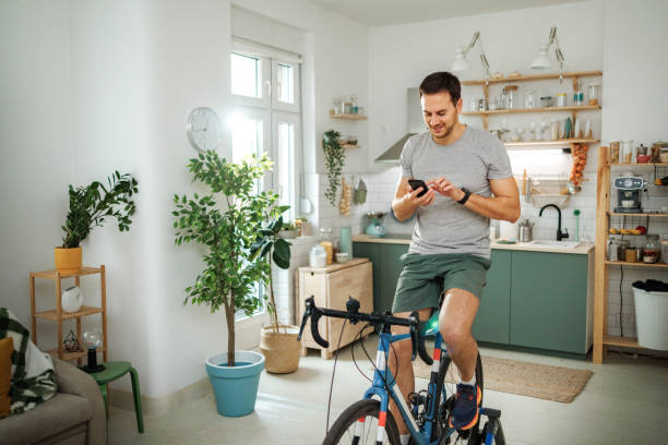 istock Young smiling handsome athletic man using smart phone while riding stationary bike at home 2149147146