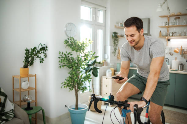 istock Young smiling handsome athletic man using smart phone while riding stationary bike at home 2149147147