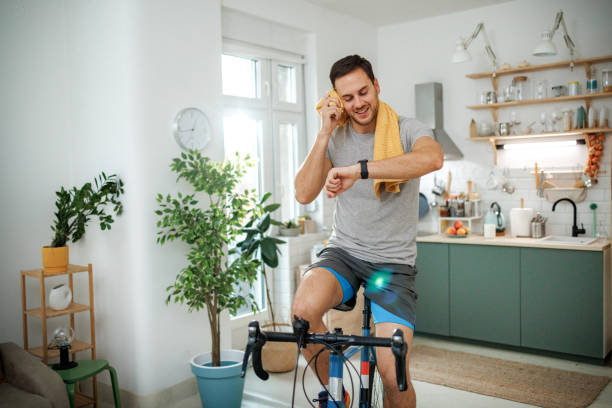 Young athletic man checking pulse on smart watch while riding bicycle in a domestic room at home
