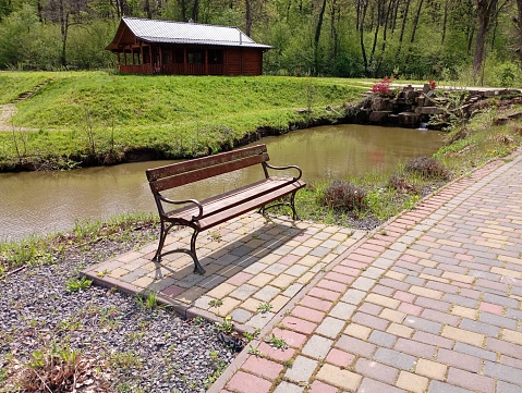 A bench in the park on a cobblestone platform near a walking path in the background of a house and a reservoir filled with water. A beautiful place to relax by the water in the park.