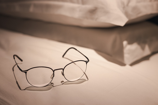 A pair of glasses is lying on a bed. The bed is unmade and the pillows are stacked on top of each other. Concept of relaxation and comfort, as the glasses are placed on a soft surface =