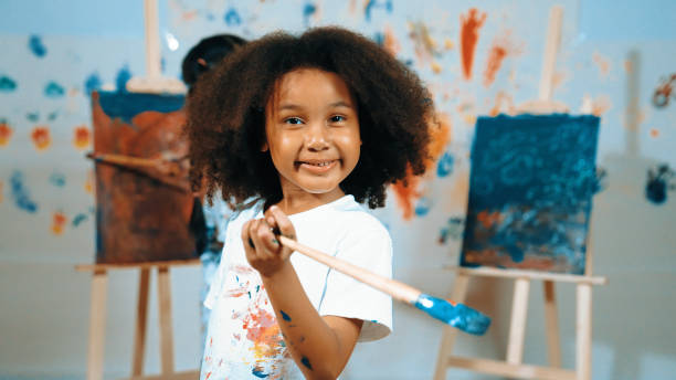 istock Cute girl holding painted brush while student drawing canvas behind. Erudition. 2149152219