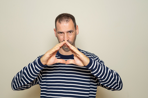 Hispanic man with a beard with his fingertips together in an expression of evil, cunning and cold planning a crime. Isolated on beige background