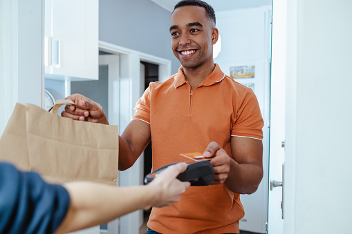 Photo of young African American man in orange t-shirt paying with credit card to delivery person
