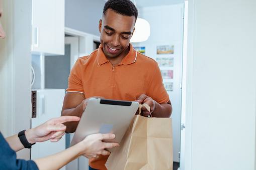 Photo of young African American man in orange t-shirt paying with credit card to delivery person