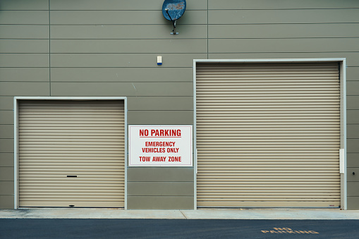 No parking, emergency vehicles only, tow away zone. Grey-green color wall with large and small garage roll-up doors. Garage for fire trucks and police vehicles.